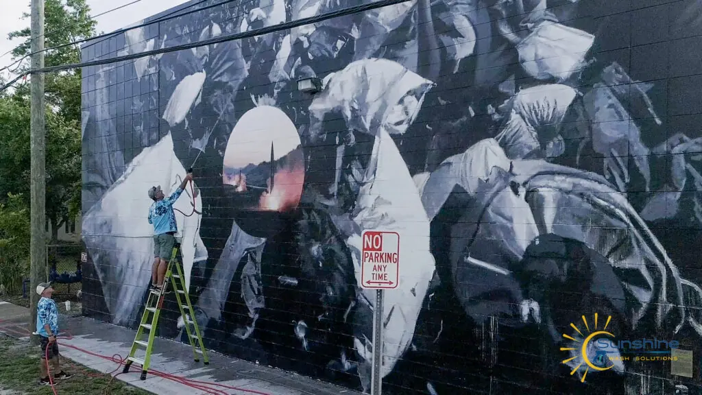 A Sunshine technician on a ladder carefully cleaning a large black-and-white mural on a building wall.